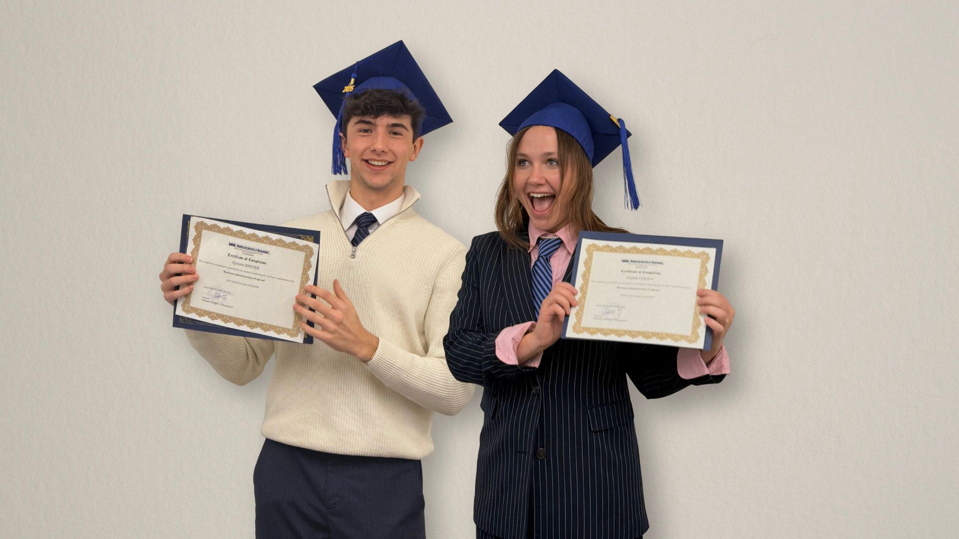 Two graduates in blue caps holding diplomas and smiling against a plain wall