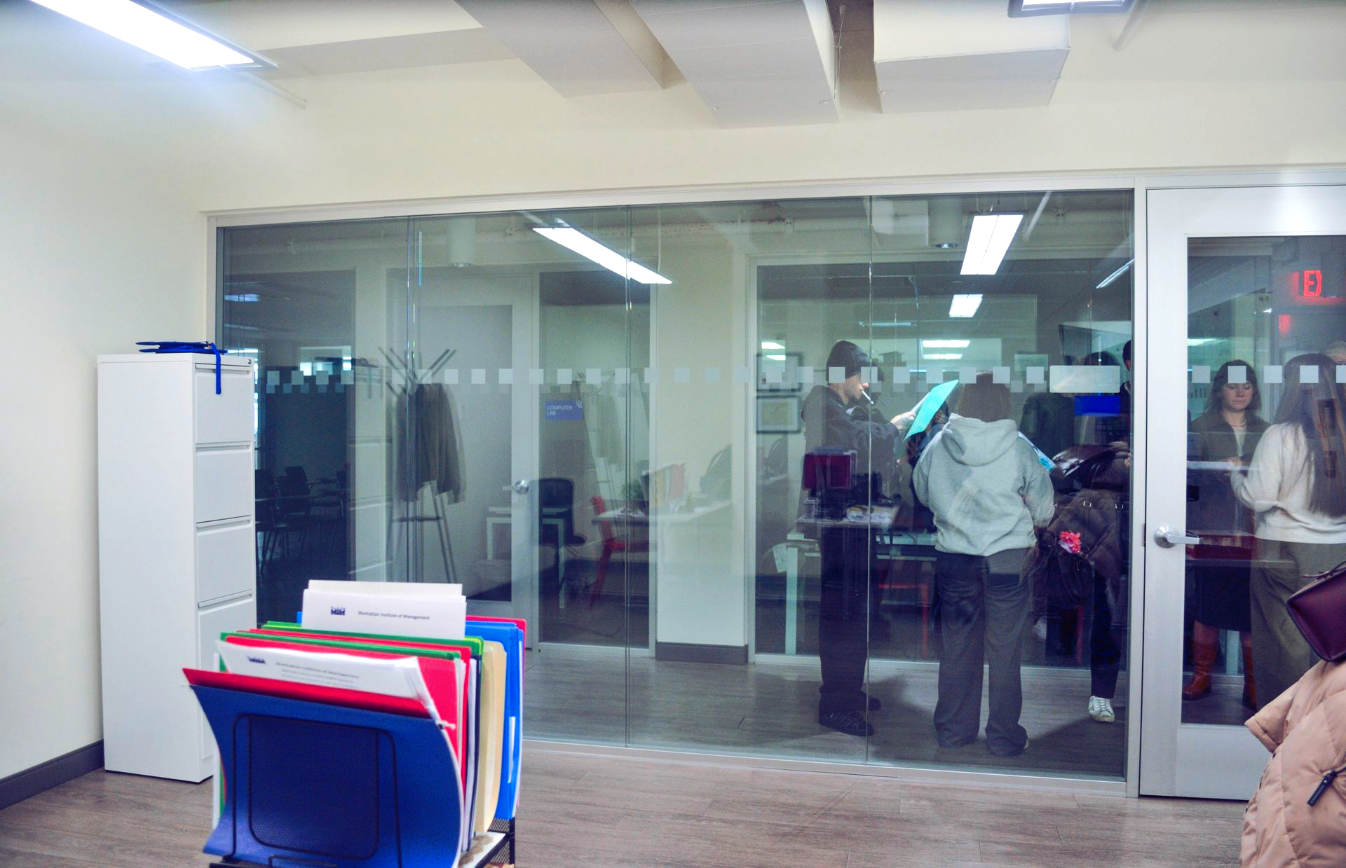 Office hallway with glass doors, people inside a meeting room, and colorful file folders in the foreground