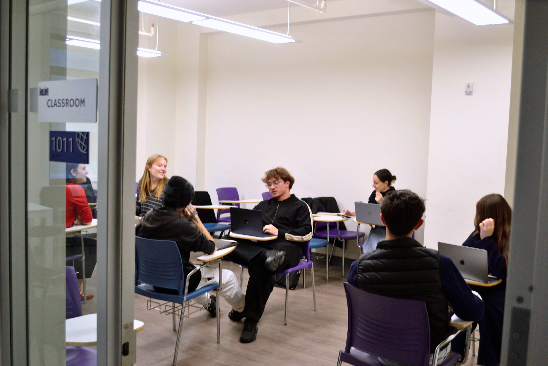 Small group meeting in a bright classroom, with people seated in a circle and talking.