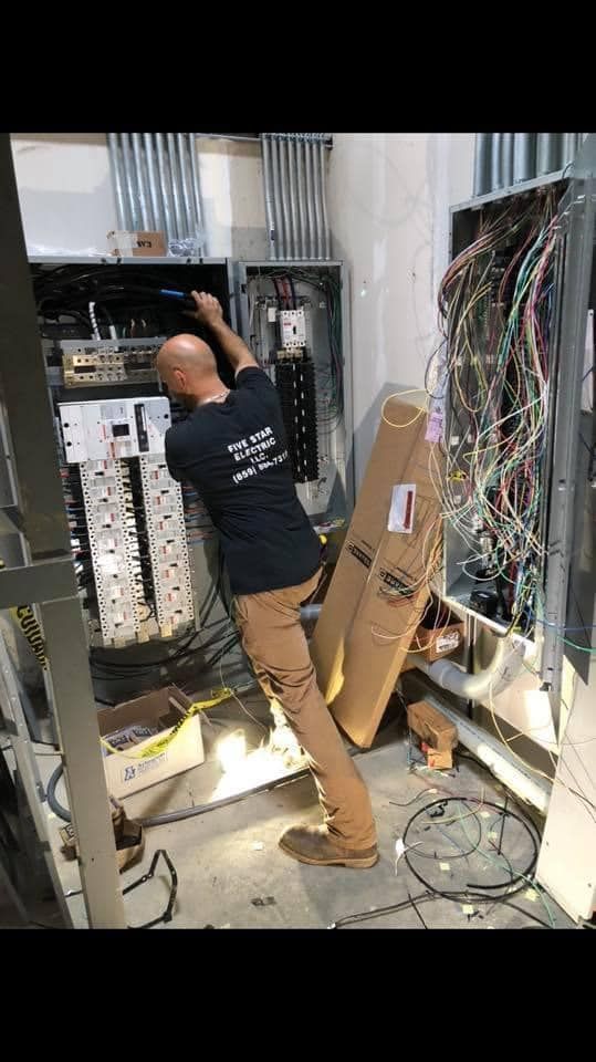 Electrician working on electrical panel, wearing black shirt and brown pants. Construction setting.