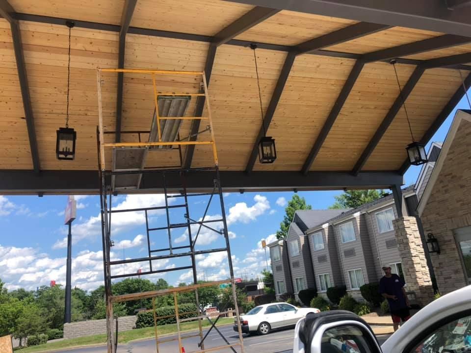 Scaffolding under a wood-paneled awning with hanging lights, in front of a building and a car.