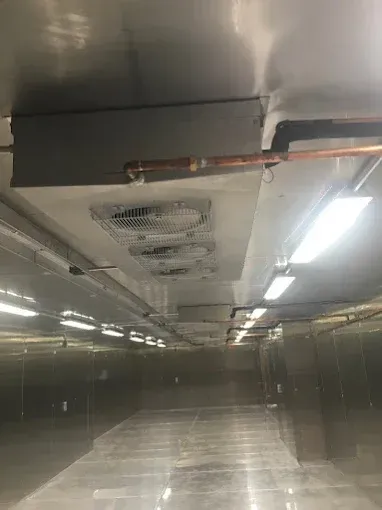 Inside a long, metallic commercial freezer with overhead fans, lights, and copper piping.