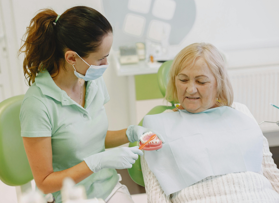 A dental professional in a mask and gloves explains a dental model to a patient in a chair.