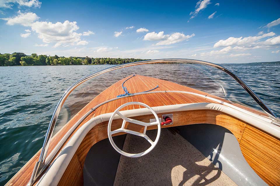 View from a small wooden boat on a lake, with a steering wheel, blue sky, and distant shoreline.