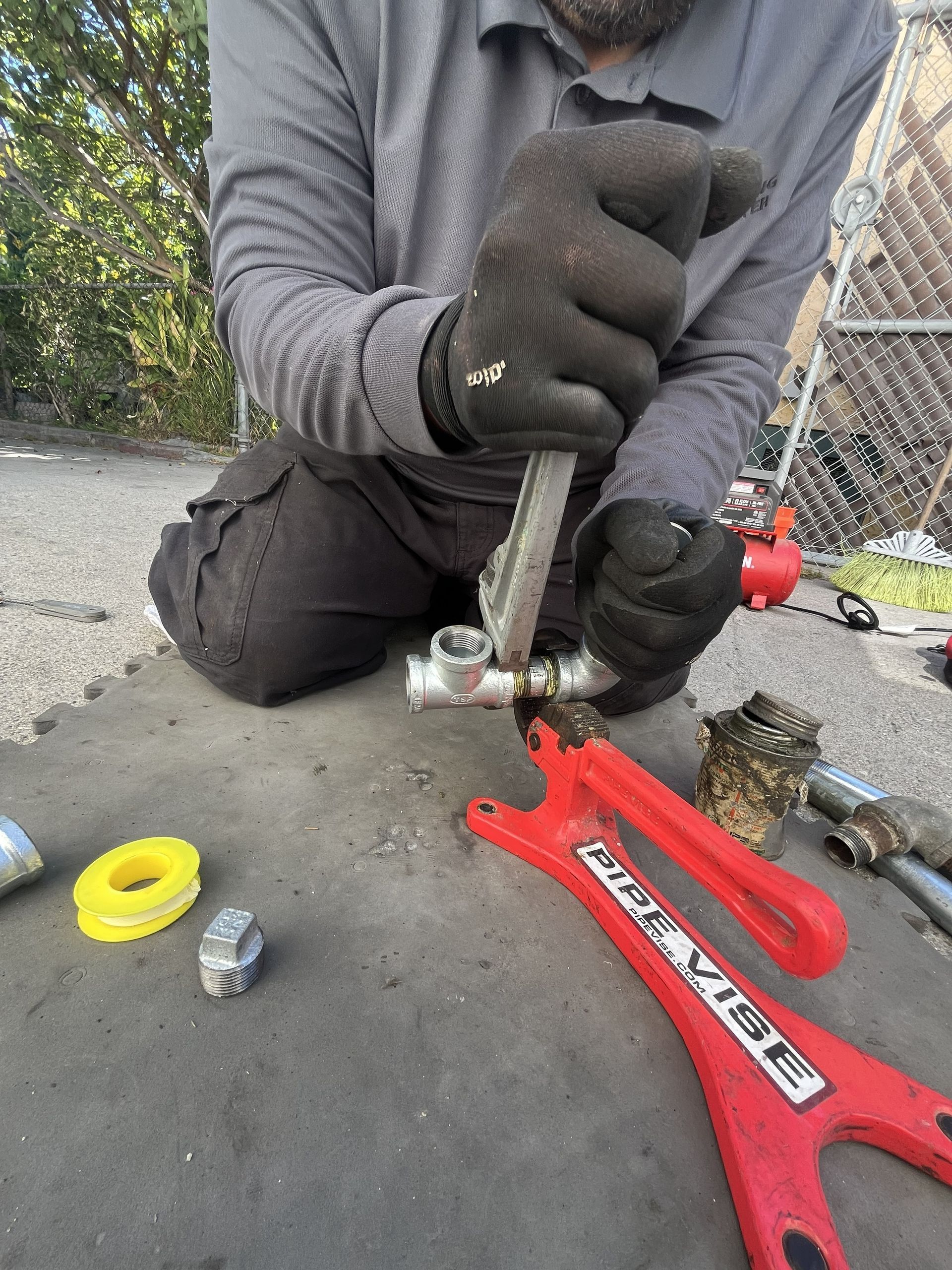 Person using a red hydraulic pipe bender outdoors on pavement, wearing black gloves and gray sleeves.