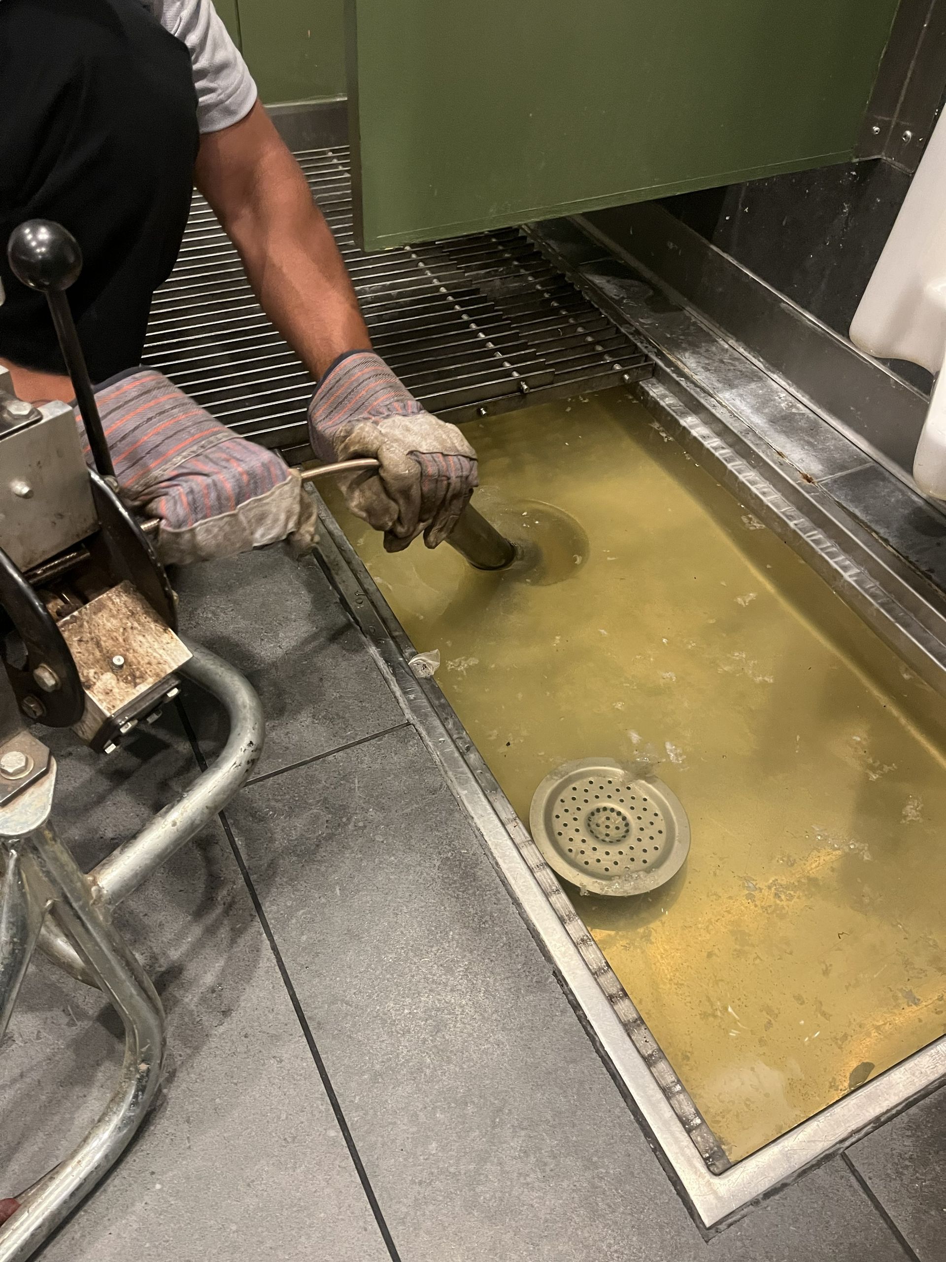 Gloved worker cleaning a greasy metal sink with yellow liquid and a drain in an industrial kitchen