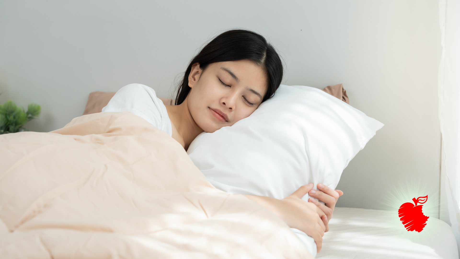 Woman sleeping in bed under a beige blanket, holding a white pillow in a bright room.