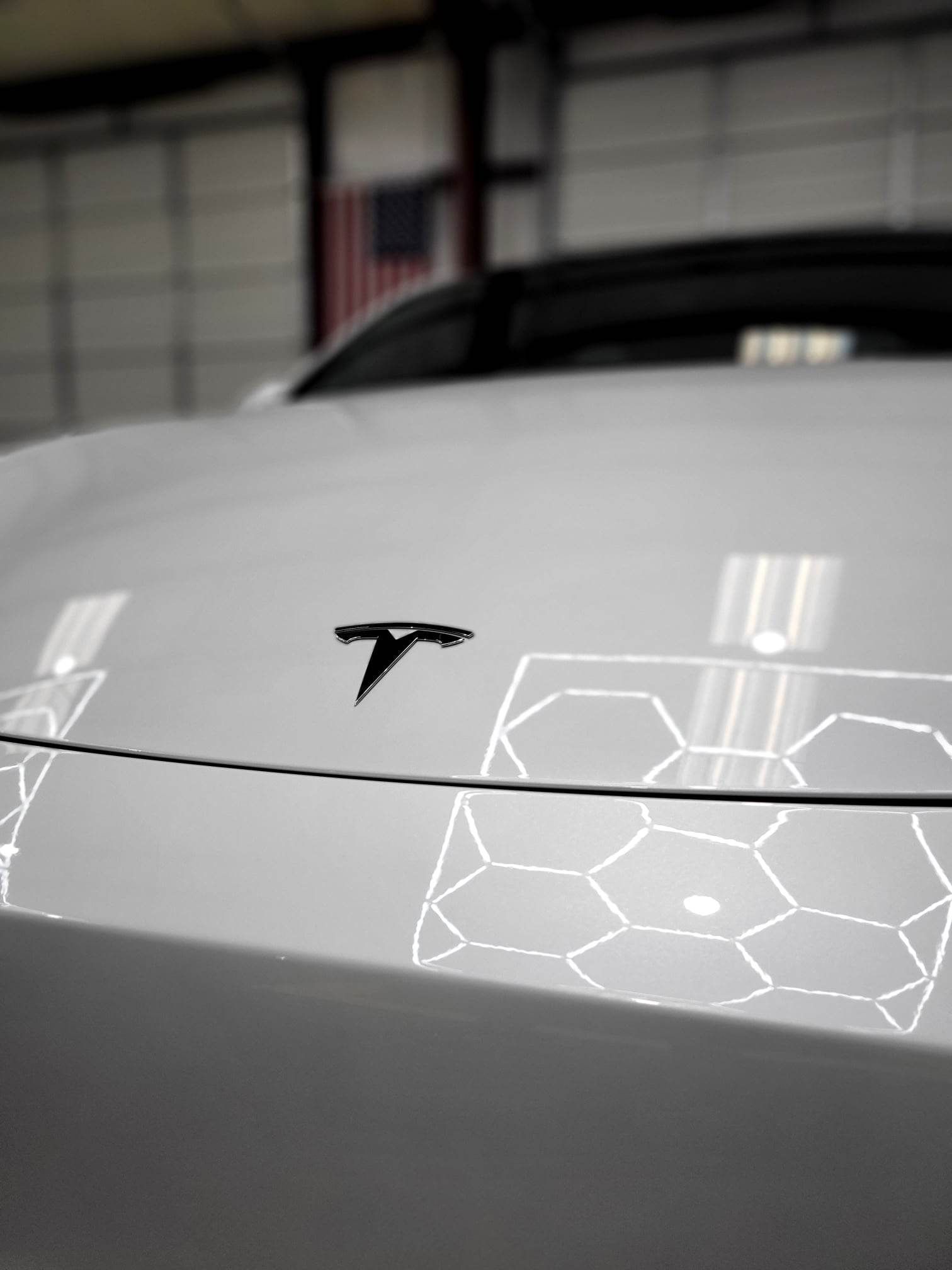 White Tesla car hood with the company logo reflecting hexagon-shaped light, setting is a garage.
