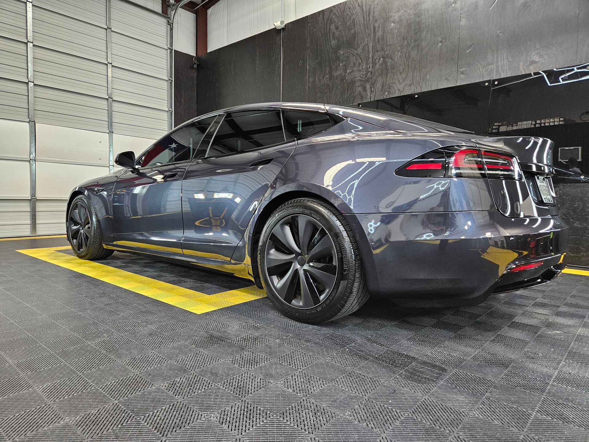 Dark gray Tesla sedan in a warehouse, parked on yellow lines near a wall.