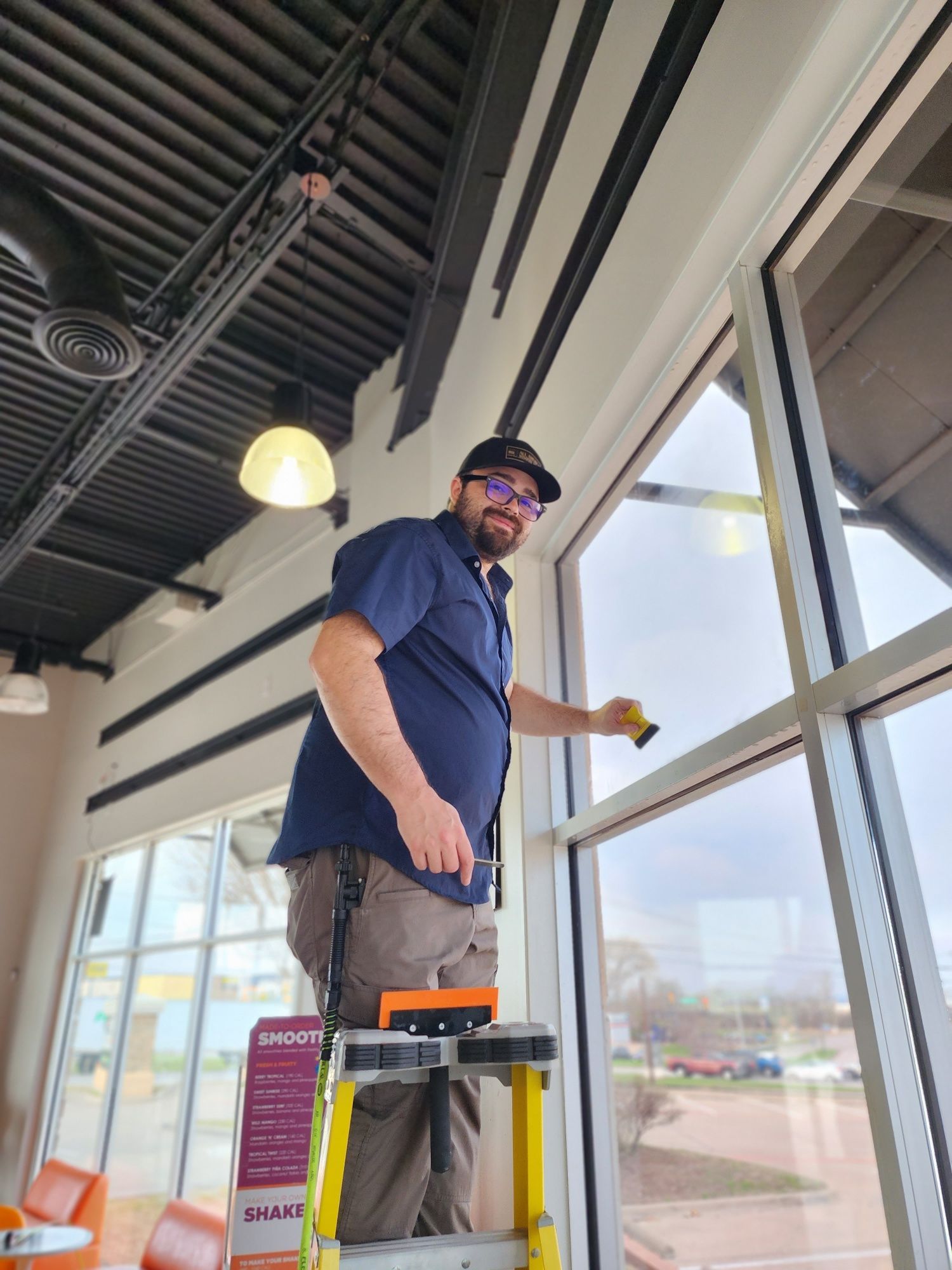 Man on a ladder cleaning a large window in a building. He smiles, holding a squeegee.