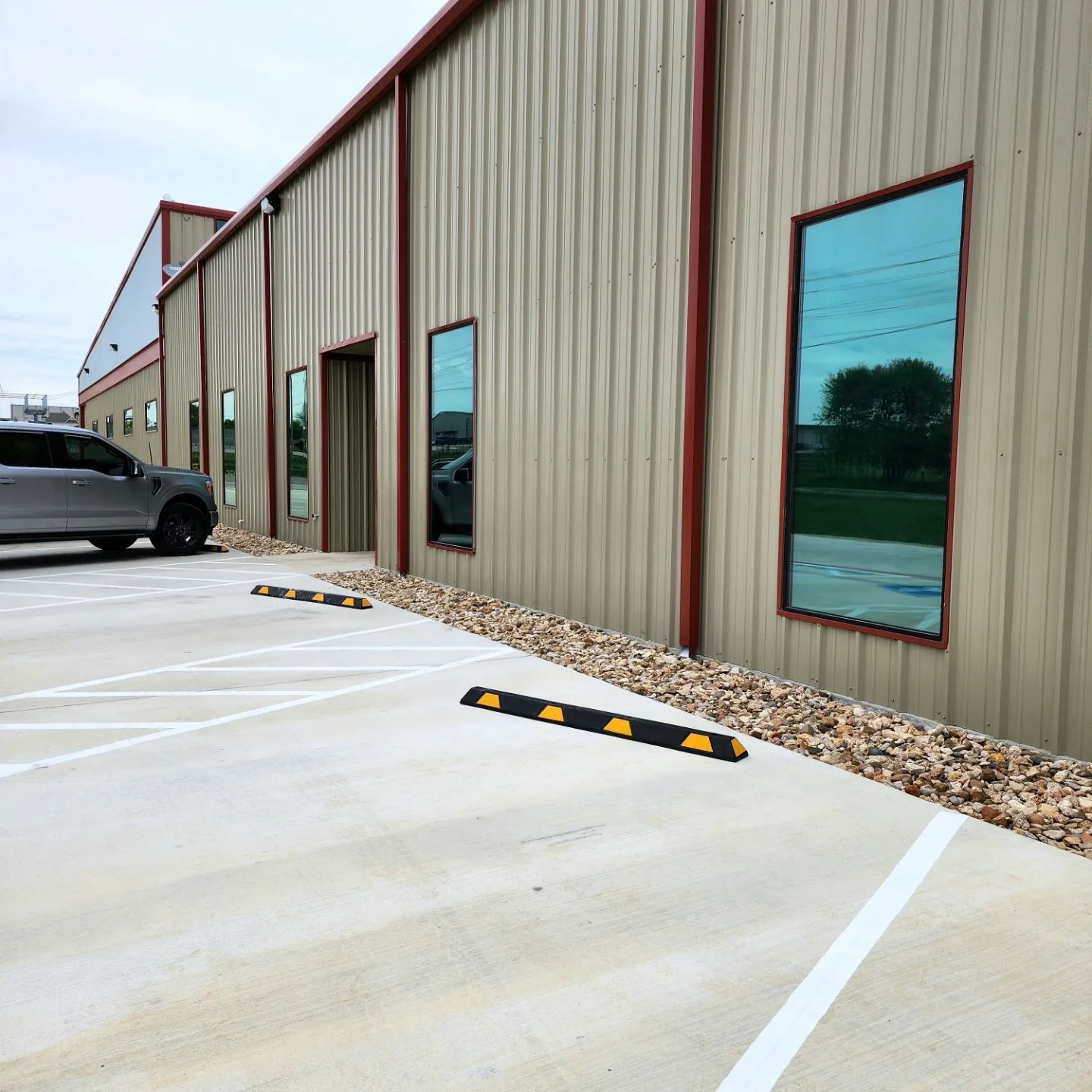 Exterior view of a building with tan siding, windows, and a parking lot. Includes a parked car and parking bumpers.