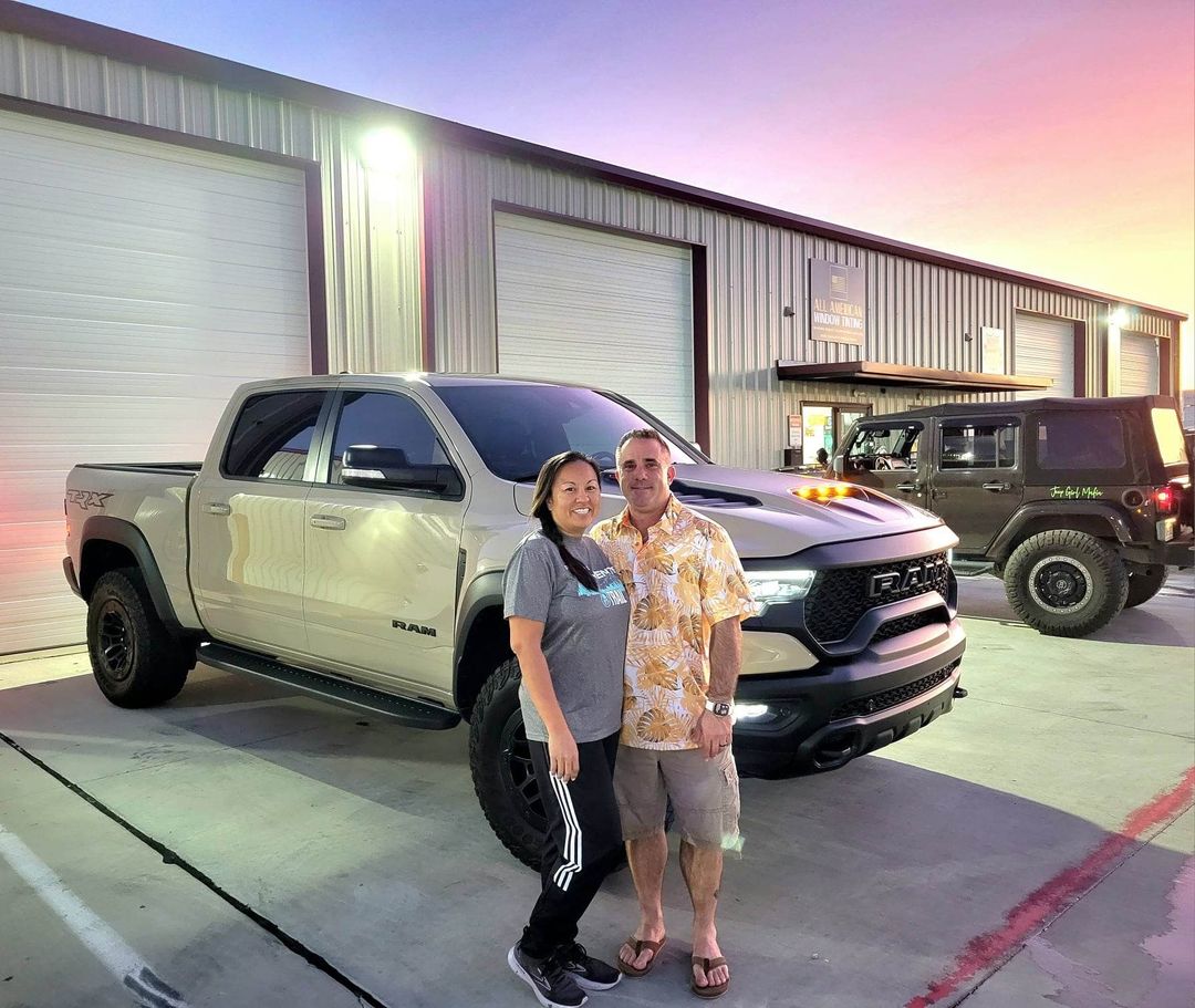 Couple posing with a tan RAM truck in front of a warehouse at sunset.