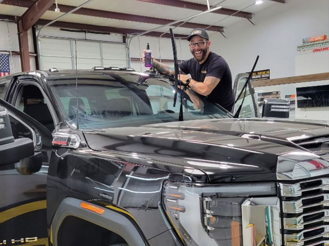 Smiling man tinting a black truck's windshield in a garage. Wipers up, holding a spray bottle.