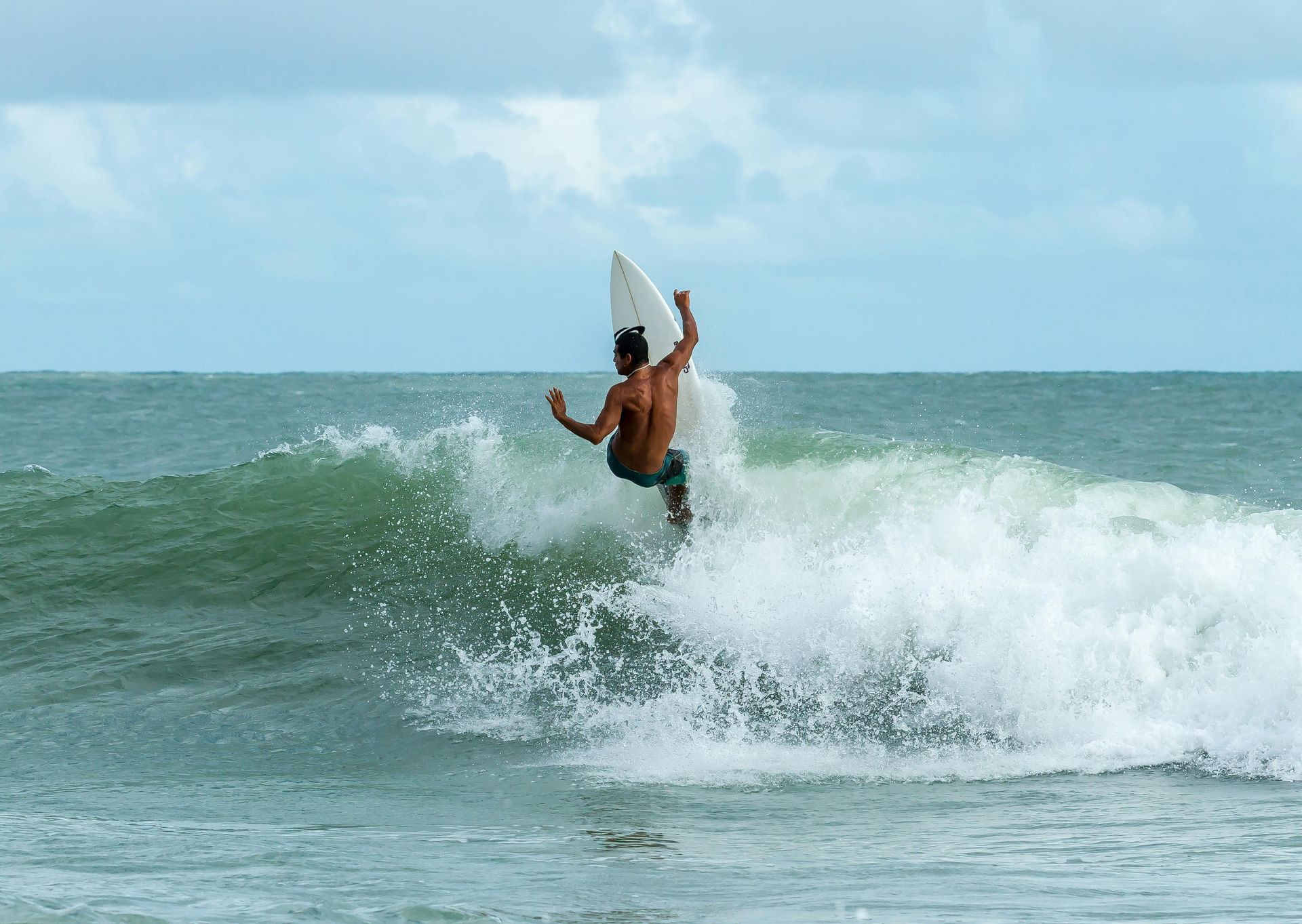 Surfer riding a wave, performing a maneuver. Ocean setting, cloudy sky. White surfboard, water splashing.