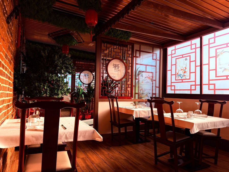Accogliente interno di un ristorante con sedie in legno, tovaglie bianche, fiori rossi e pannelli decorativi in stile cinese.