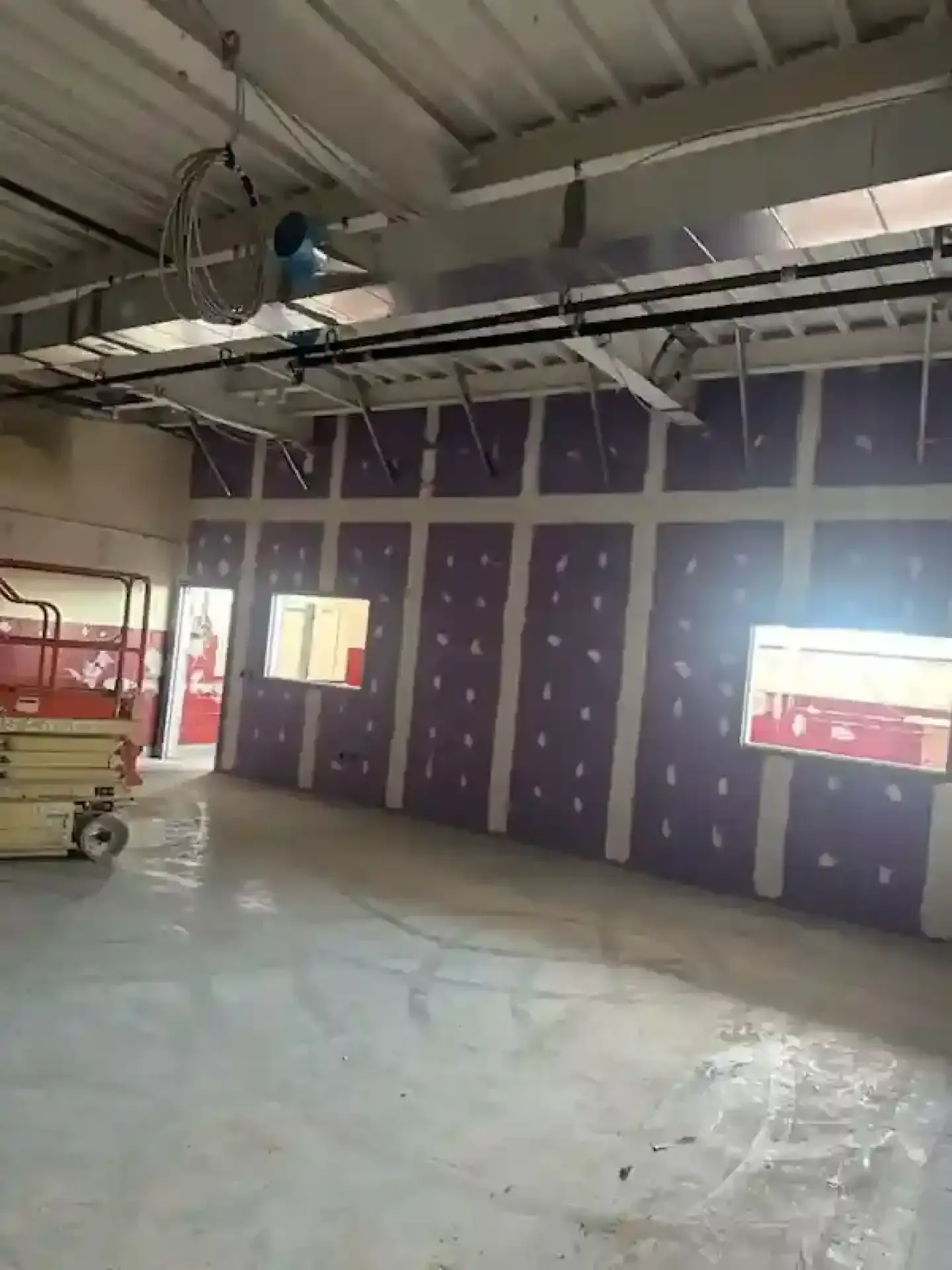 Interior of a construction site with purple wall panels, exposed ceiling, and a scissor lift on the left.