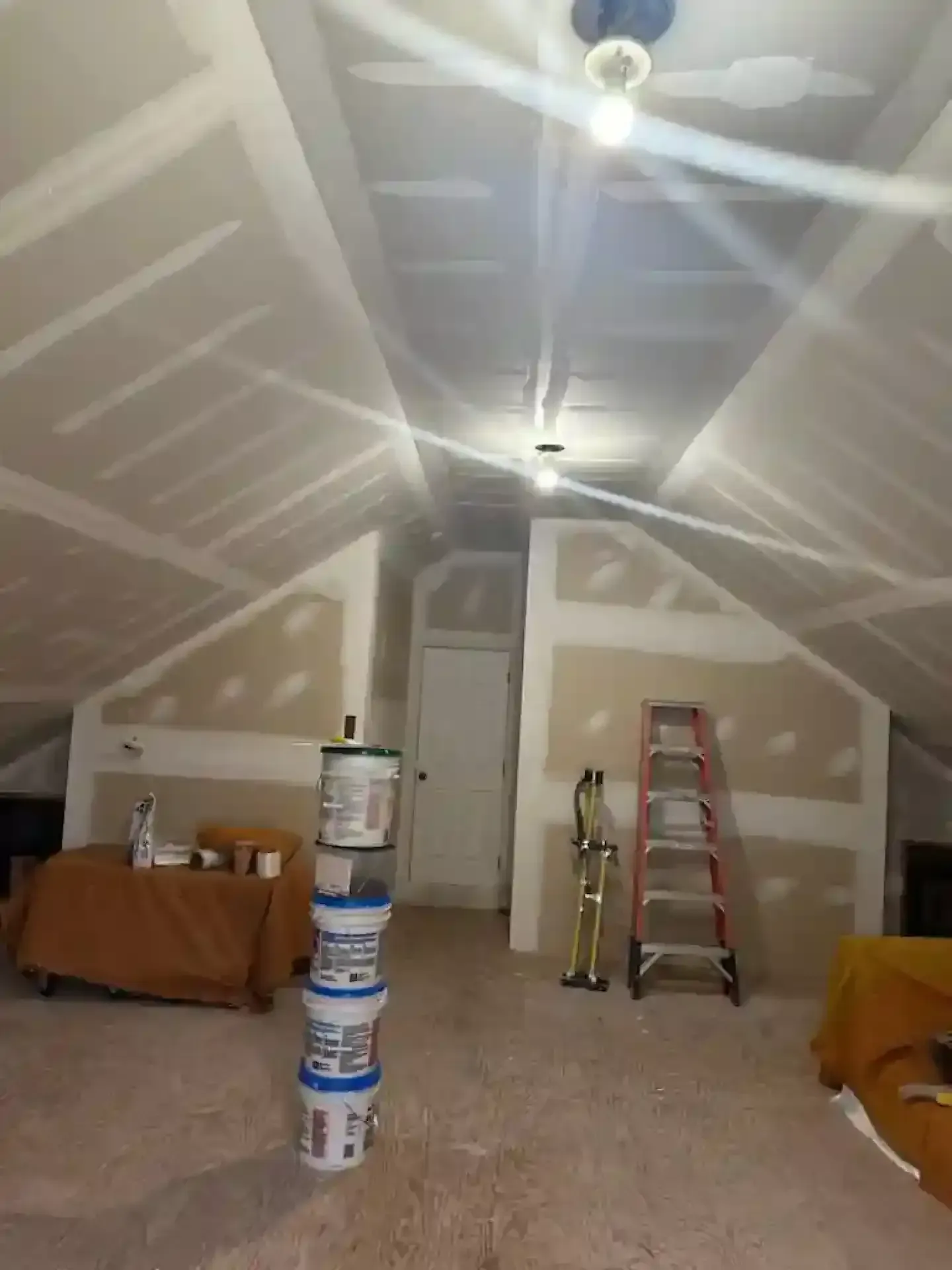 Unfinished room with drywall, ladder, stacked paint buckets, and covered furniture under bright ceiling lights