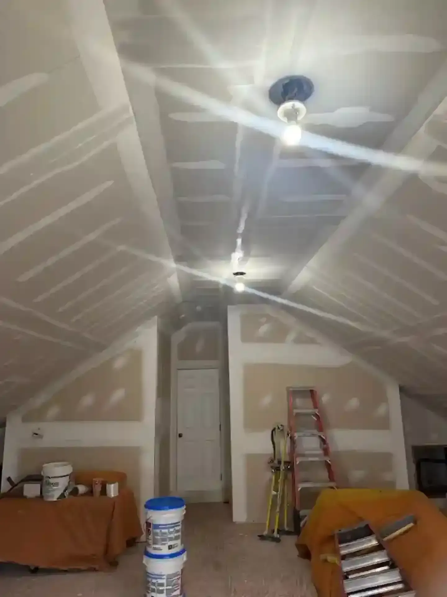 Unfinished attic room with drywall, two hanging lights, ladders, and paint supplies.