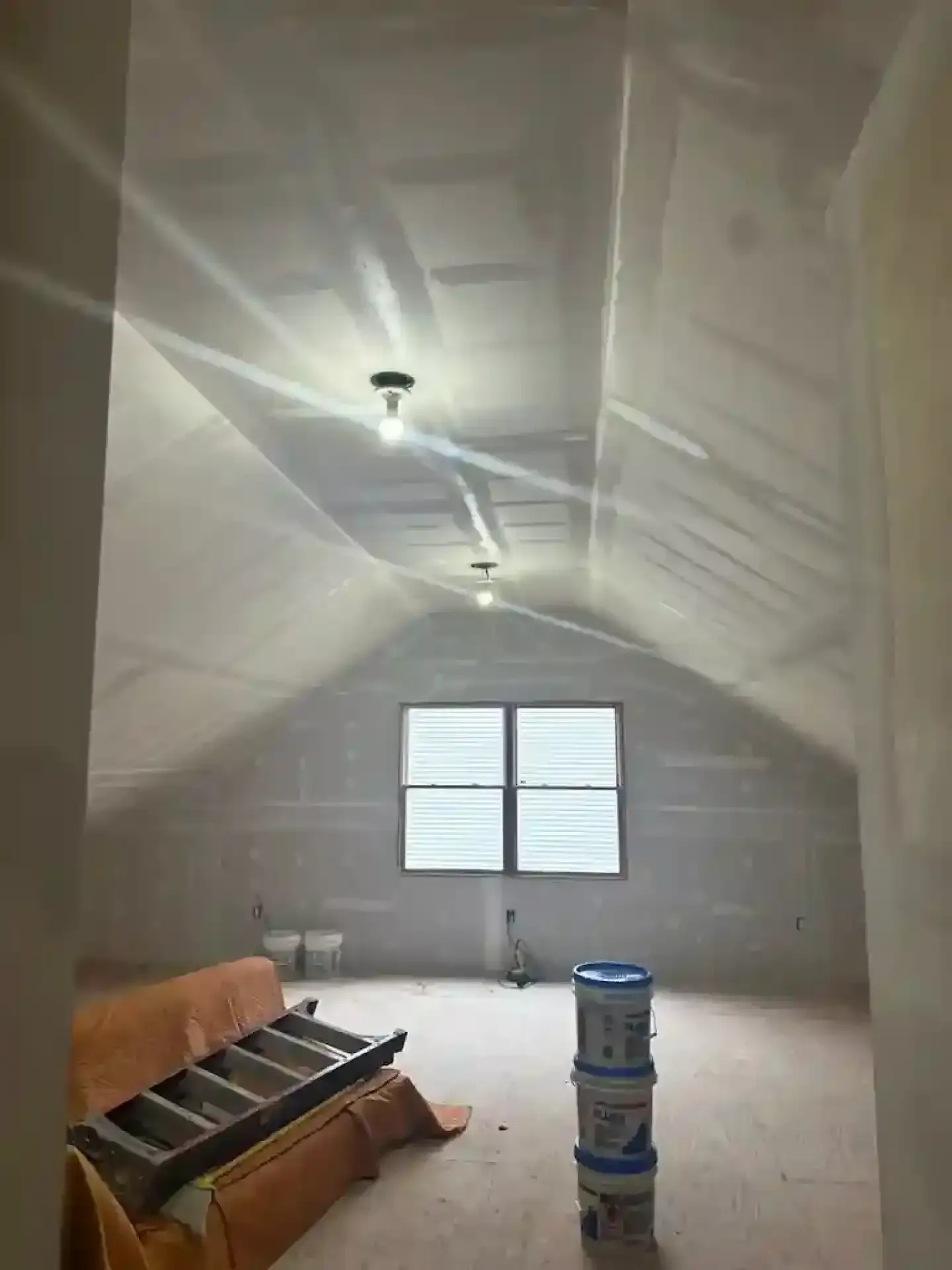 Unfinished attic room with sloped ceiling, window, exposed lights, couch, and paint buckets on the floor