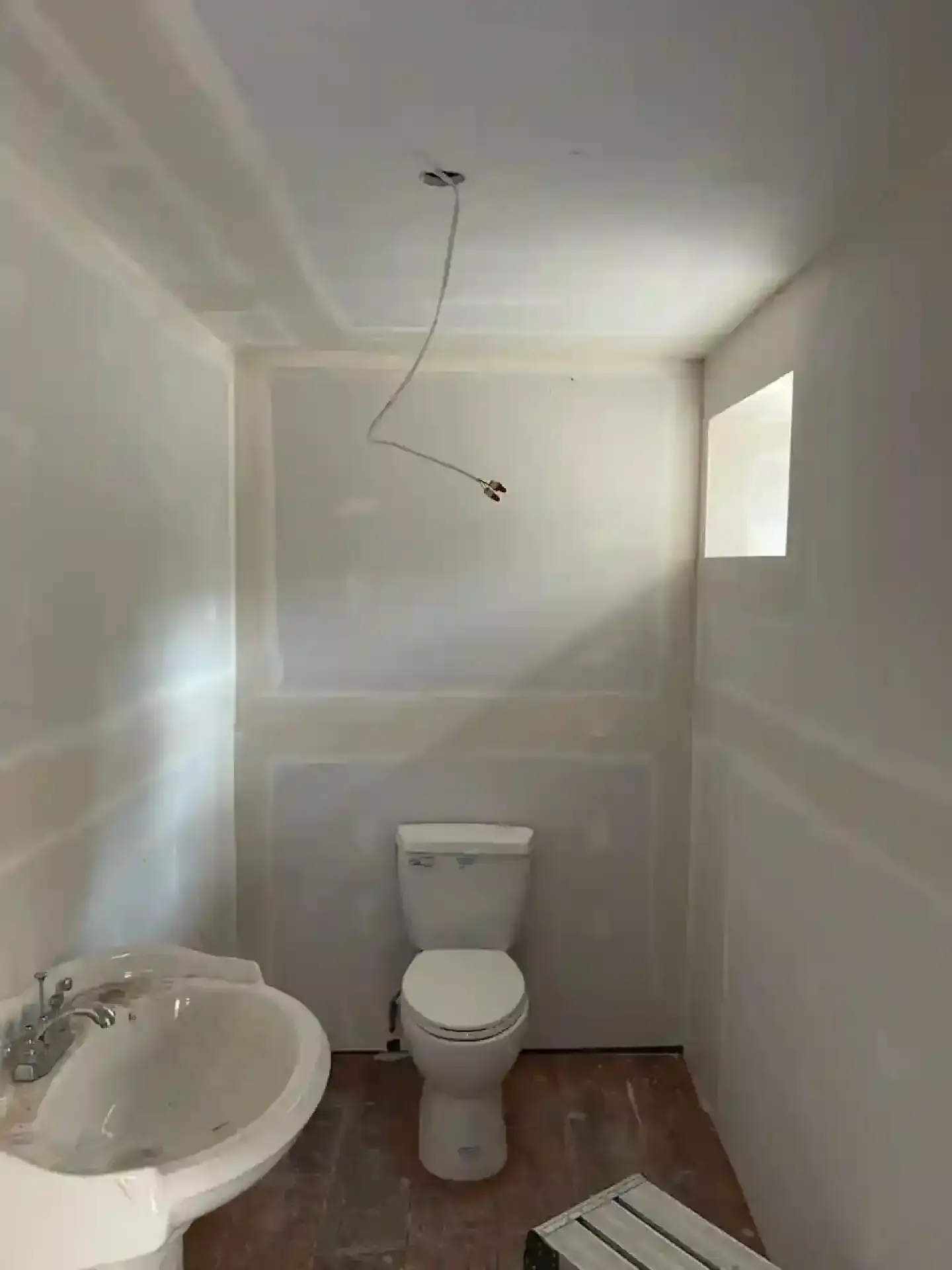 Small unfinished bathroom with sink, toilet, exposed ceiling light wiring, and a small window.