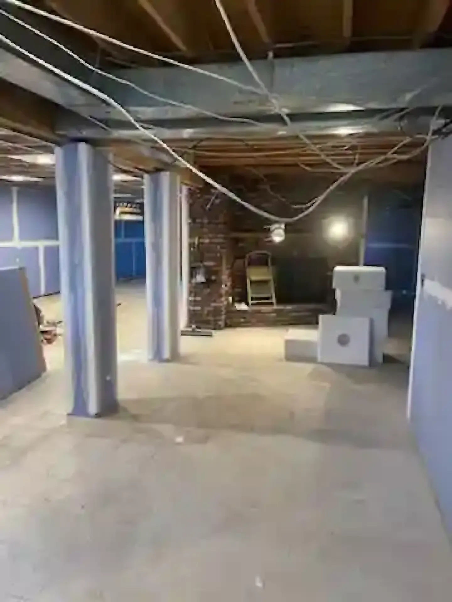 Basement with exposed ceiling joists, concrete floor, metal support poles, and stacked boxes near a brick wall