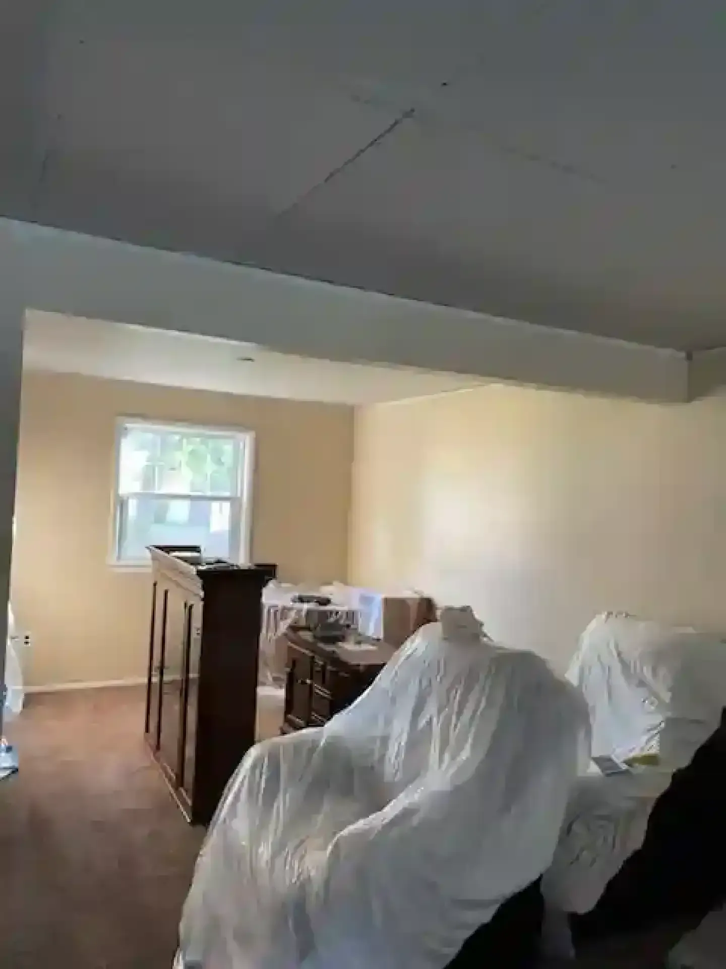 Partially furnished room with covered furniture, a wooden dresser, and a small window in beige walls