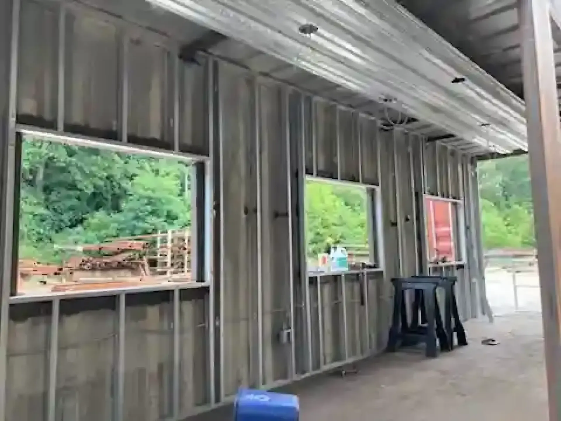 Interior of an unfinished metal building with open windows, a black table, and green trees outside