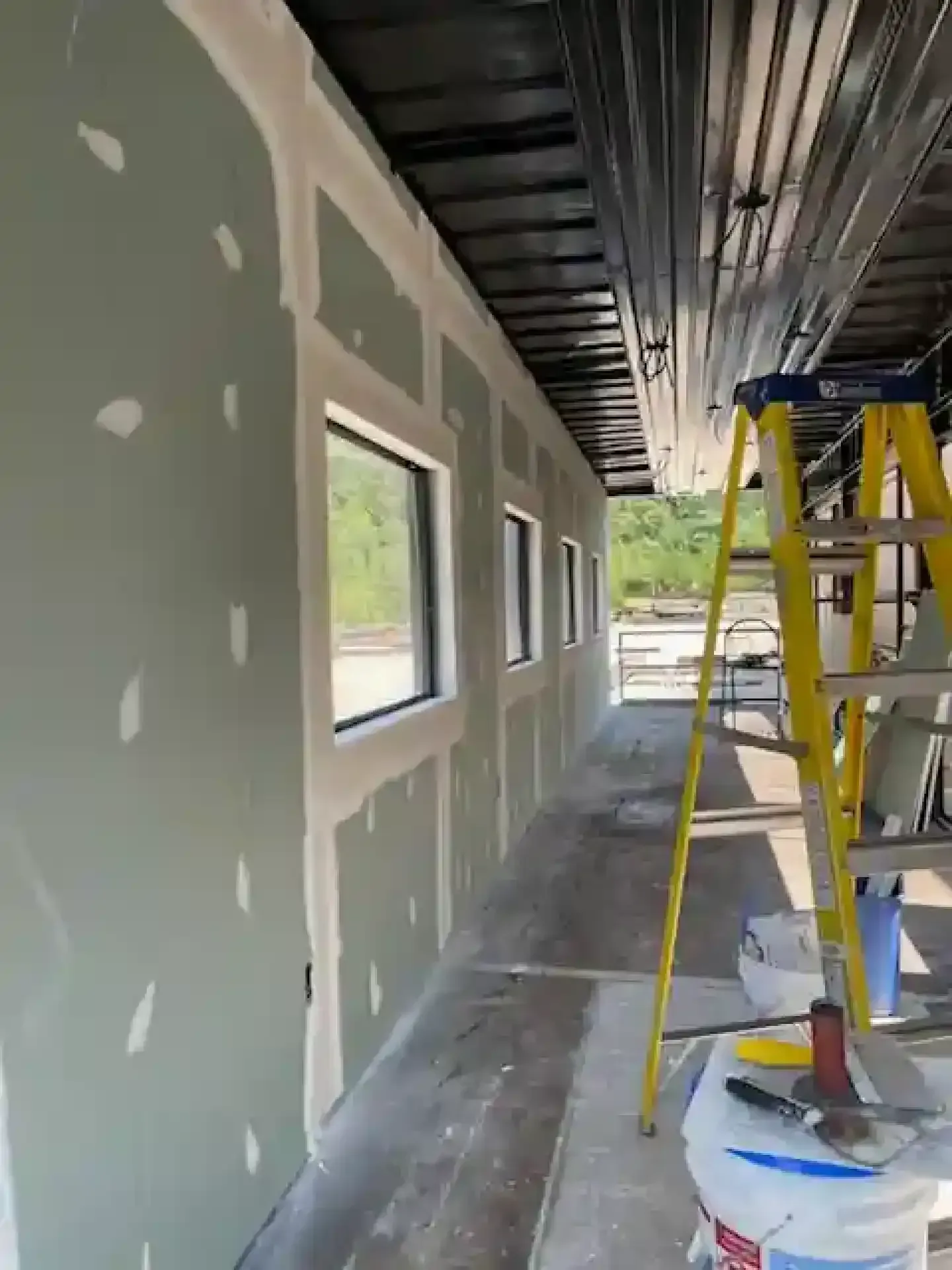 Narrow hallway under construction with gray drywall, windows, a ladder, and painting supplies