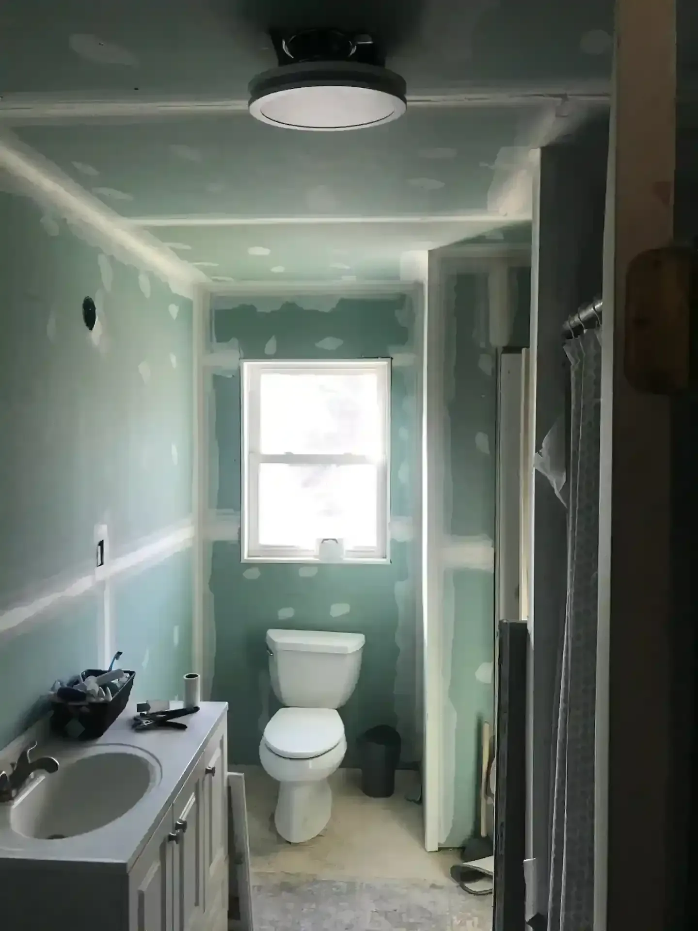 Small bathroom under renovation with mint green walls, toilet, sink, and window.
