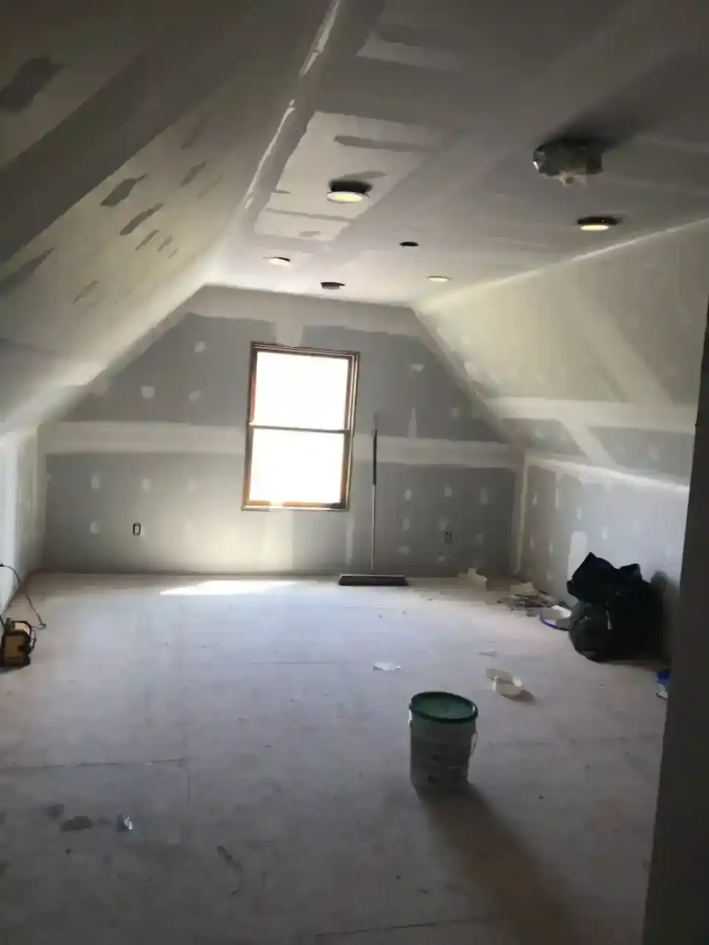 Unfinished attic room with drywall, sloped ceiling, central window, and a paint bucket on the floor