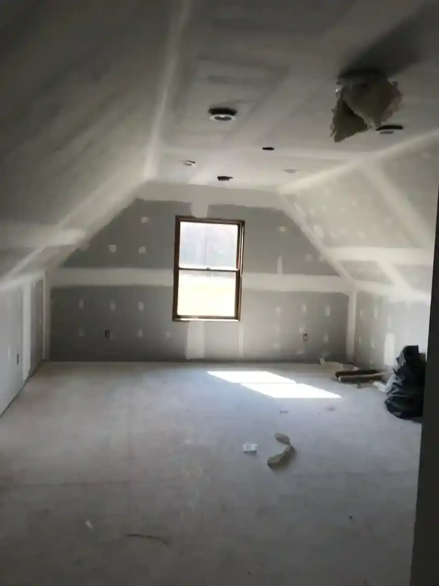 Unfinished attic room with sloped ceiling, drywall patches, a small window, and construction debris on the floor