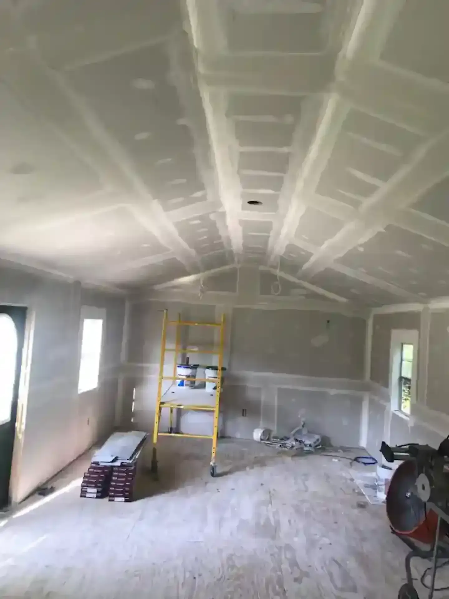 Unfinished room with drywall ceiling, windows, ladders, and construction supplies spread across the floor.