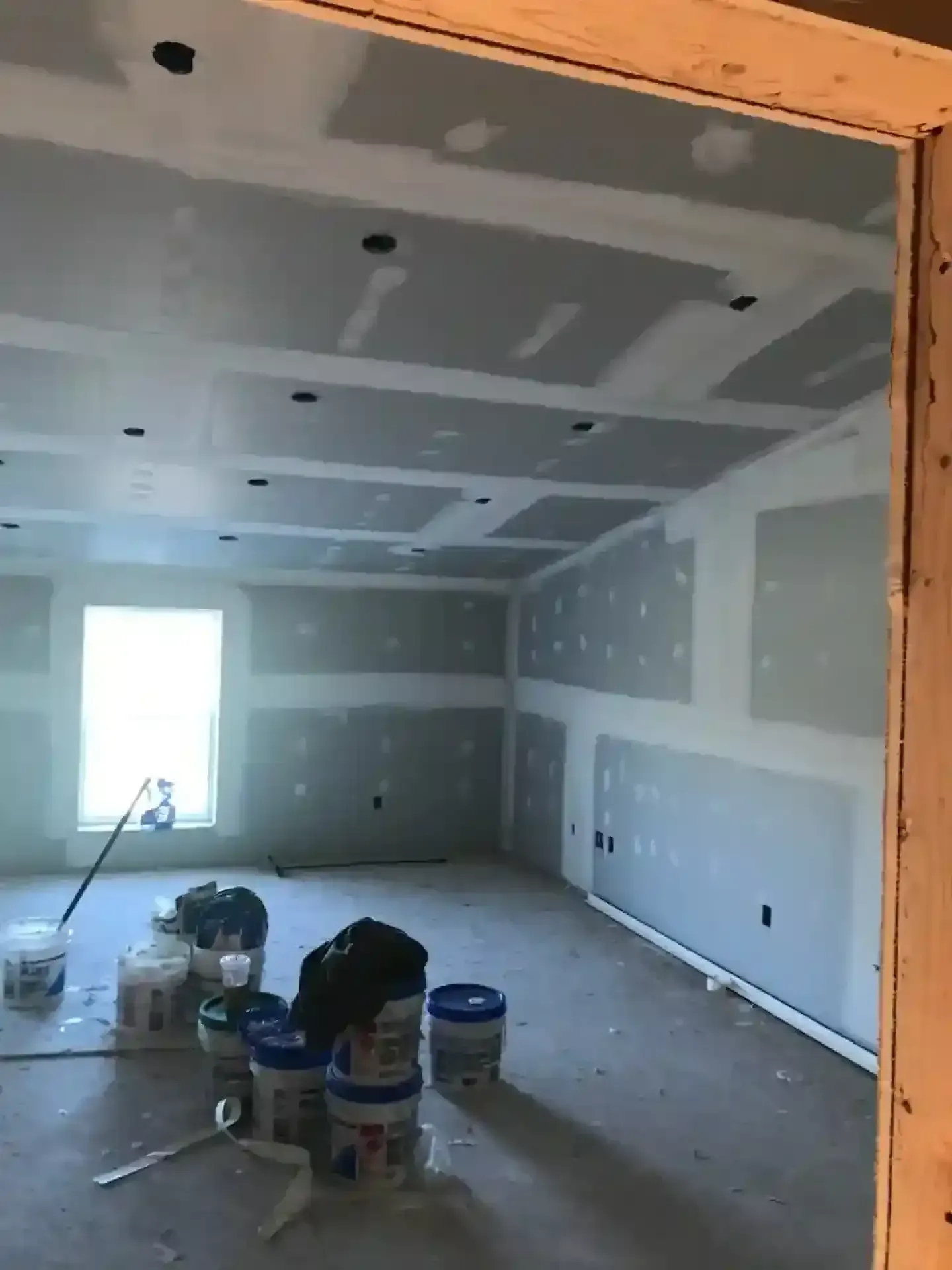 Unfinished room with drywall, paint buckets, tools, and a bright window at the far end.