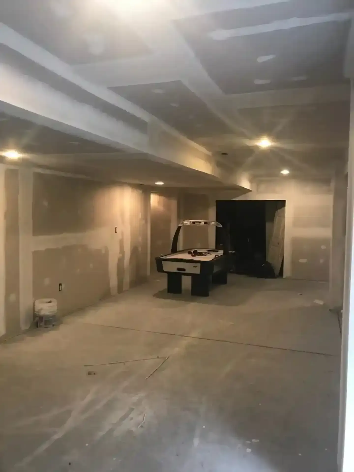 Empty unfinished room with drywall dust, ceiling lights, and a work table in the center.