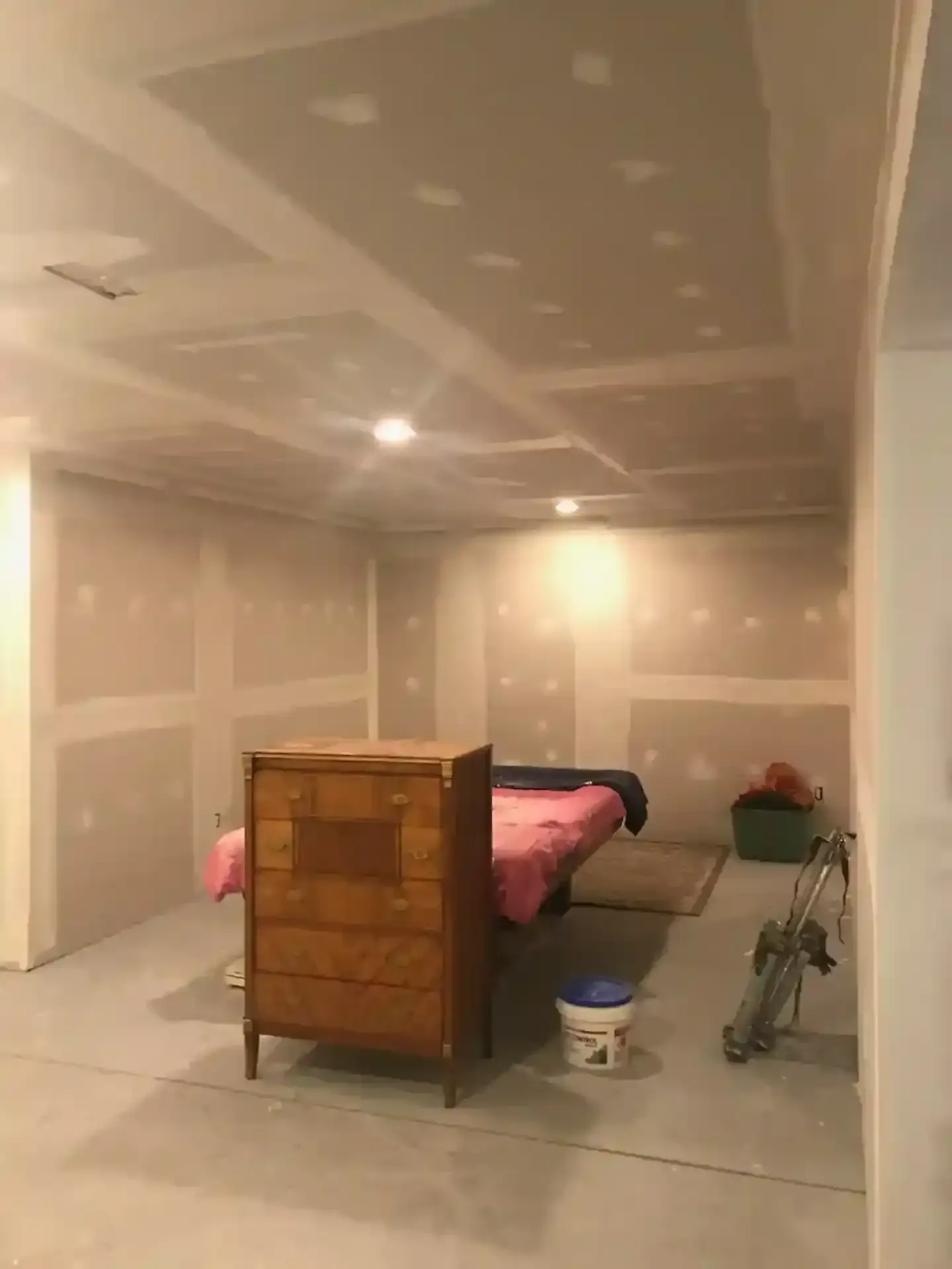 Unfinished bedroom with drywall, bed, dresser, and ceiling lights; a bucket sits on the floor.