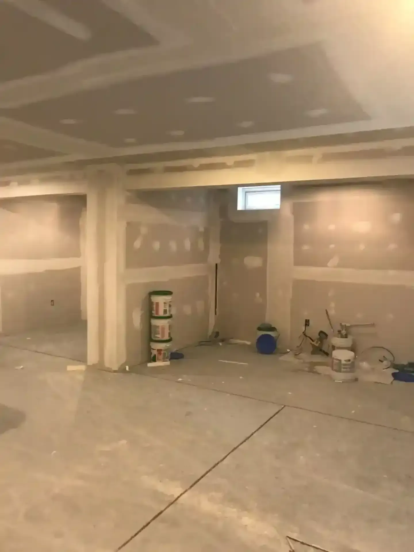 Unfinished drywall room with taped seams, dusty floor, buckets, and tools in a construction site.