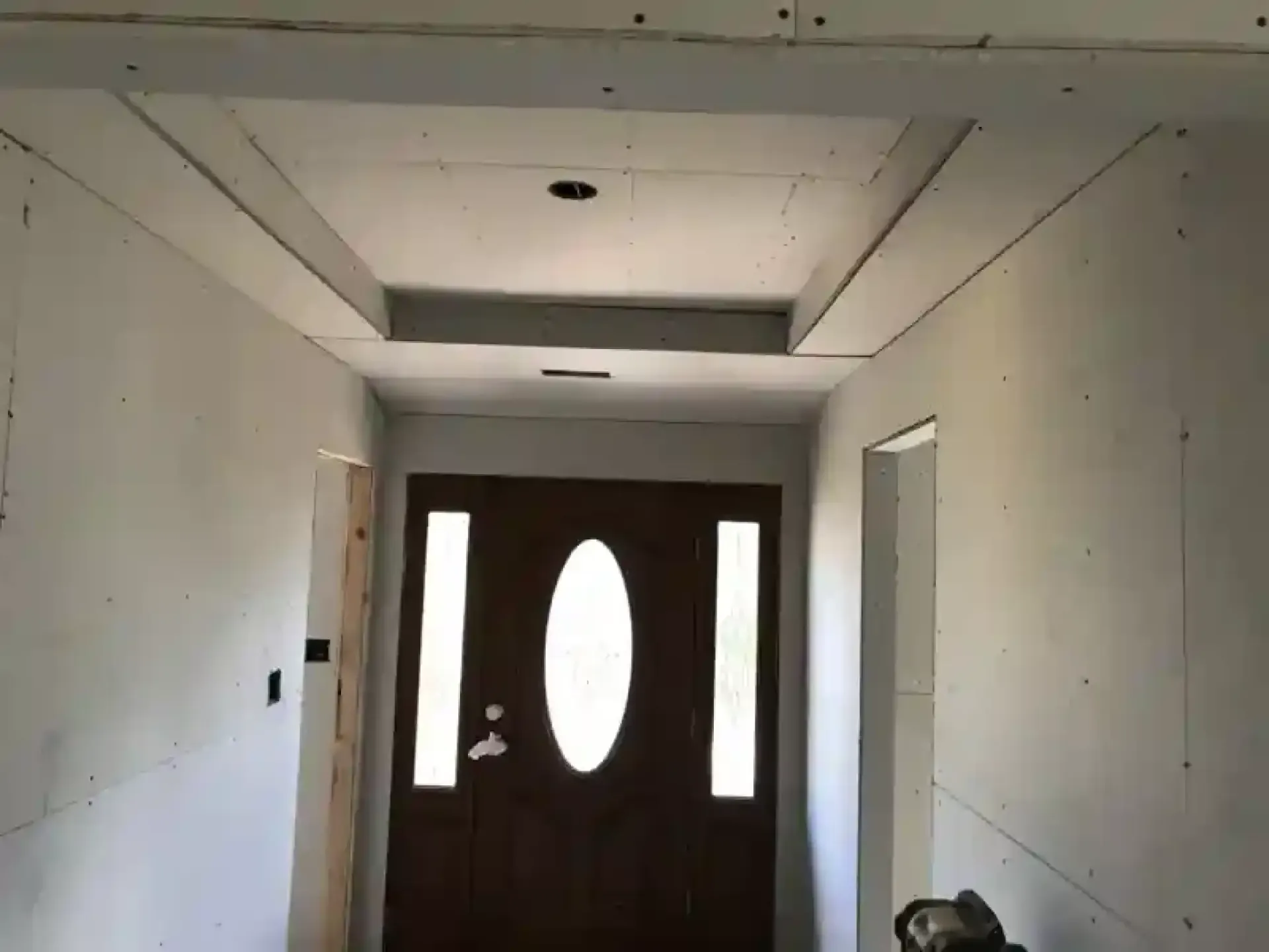 Unfinished hallway with drywall leading to a front door featuring oval and sidelights