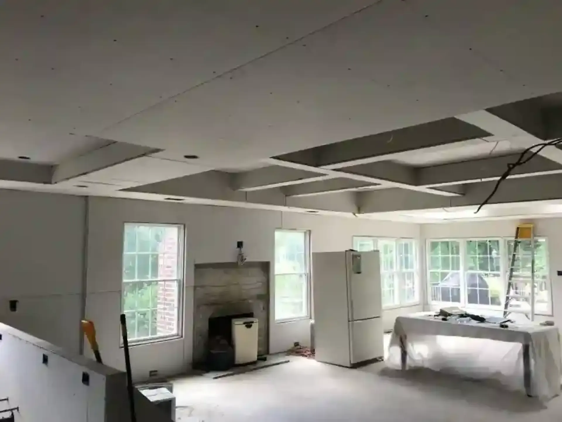 Unfinished room with white walls, exposed ceiling, windows, a fireplace, and covered furniture near the right wall