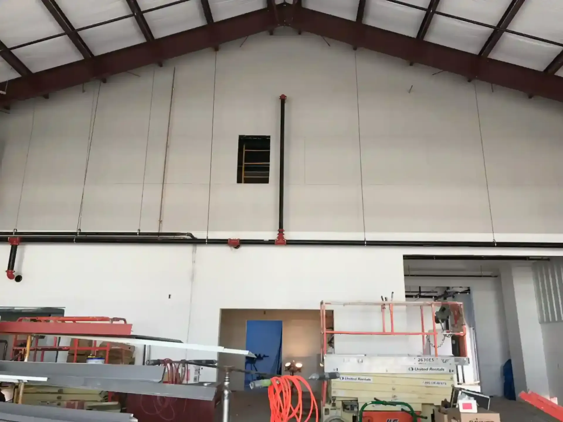 Interior of a warehouse with a high white wall, exposed beams, and stacked materials along the floor.