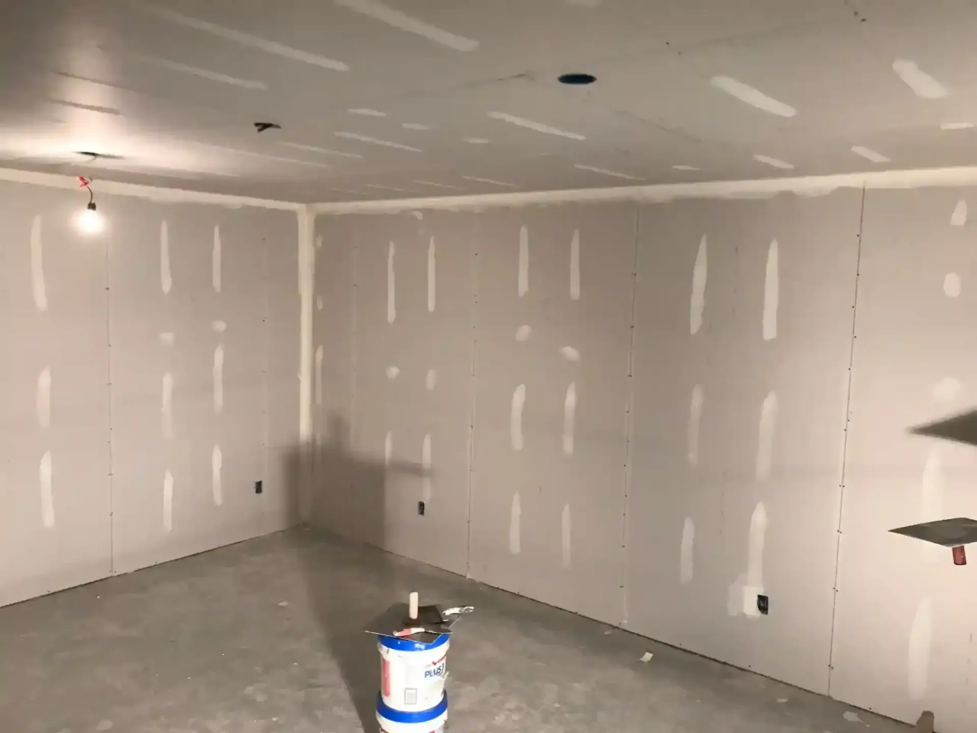 Empty drywall room under construction with white taped seams and a bucket on the concrete floor