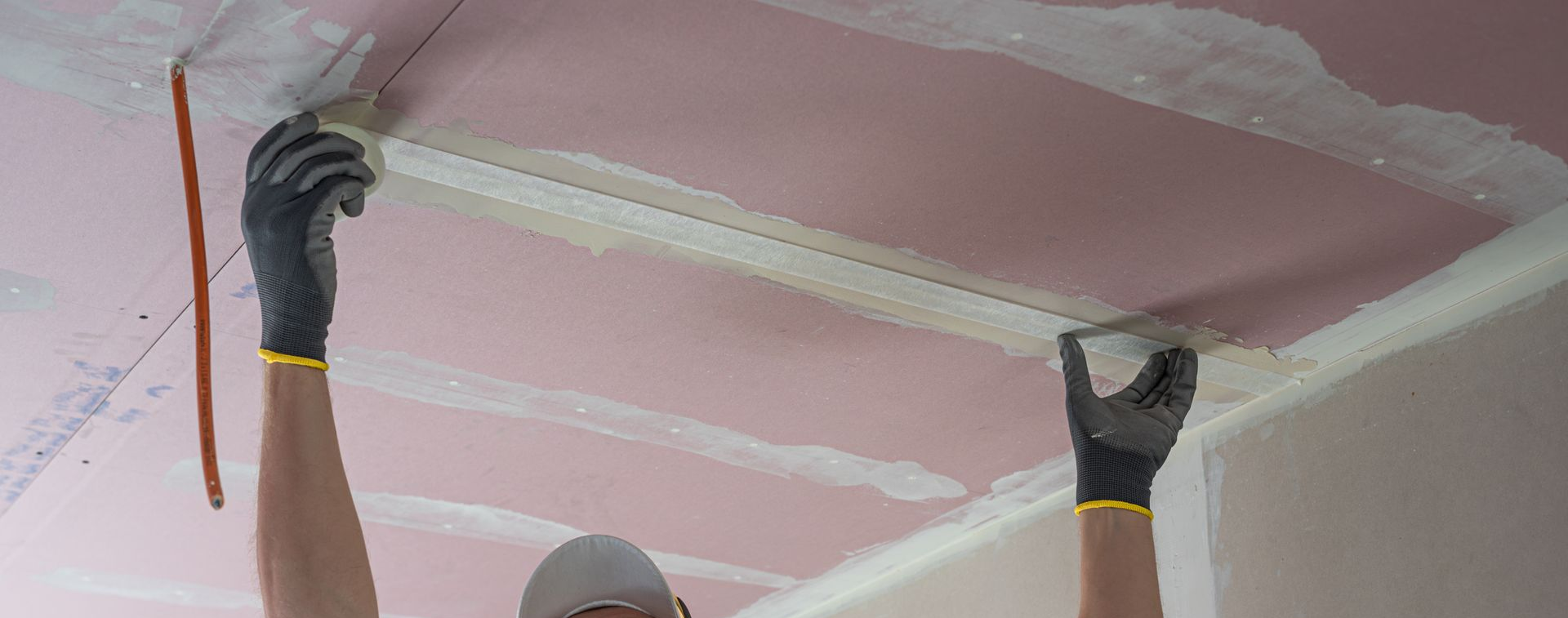Gloved hands smoothing white trim along a pink ceiling during home painting/renovation