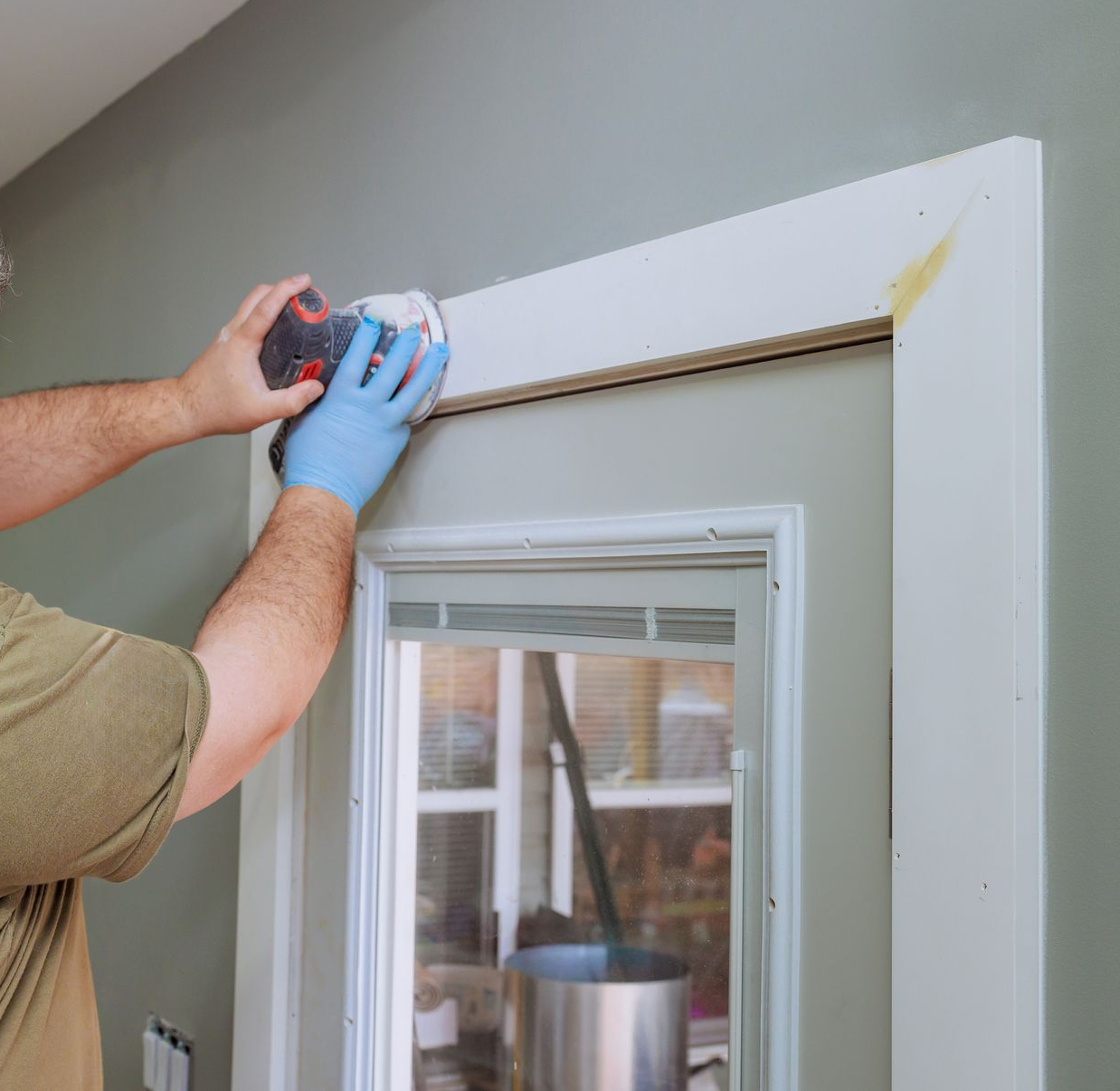Person installing trim above a window with a drill, wearing blue gloves.