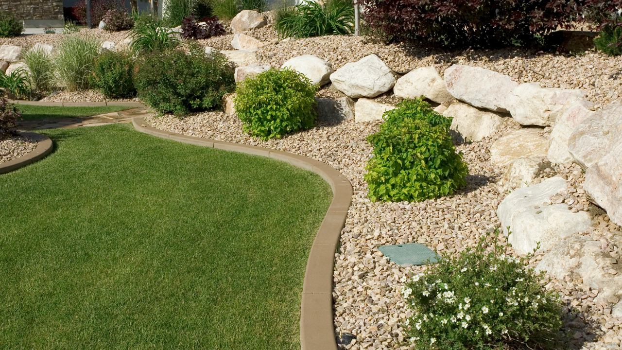 A lush green lawn curves along a tan concrete border, flanked by decorative rocks, shrubs, and a low stone retaining wall.