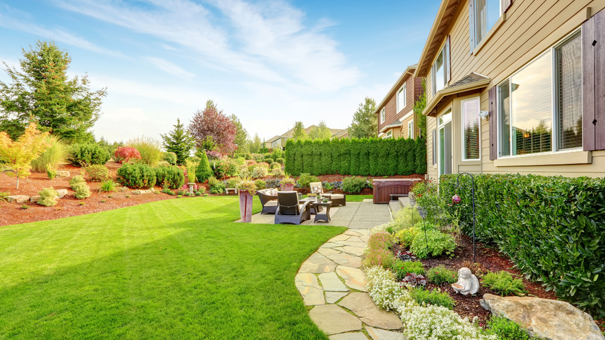 A residential backyard with a green lawn, a flagstone path, garden beds, and a patio area with outdoor furniture.