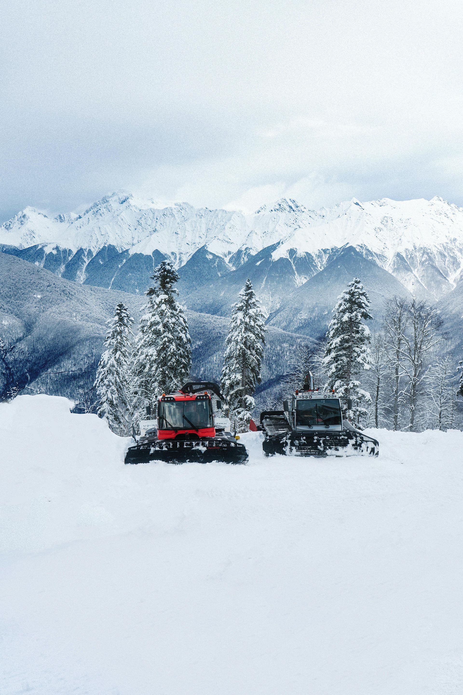 Two snow grooming machines parked on a snowy mountain slope with pine trees and snow-capped peaks in the background.