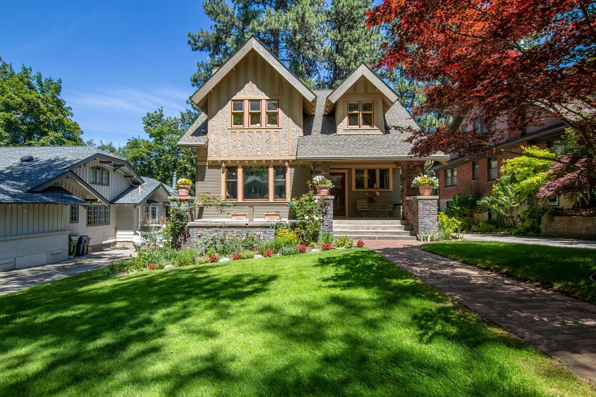 A two-story tan craftsman-style home with a gabled roof, stone accents, and a lush front lawn under a clear blue sky.