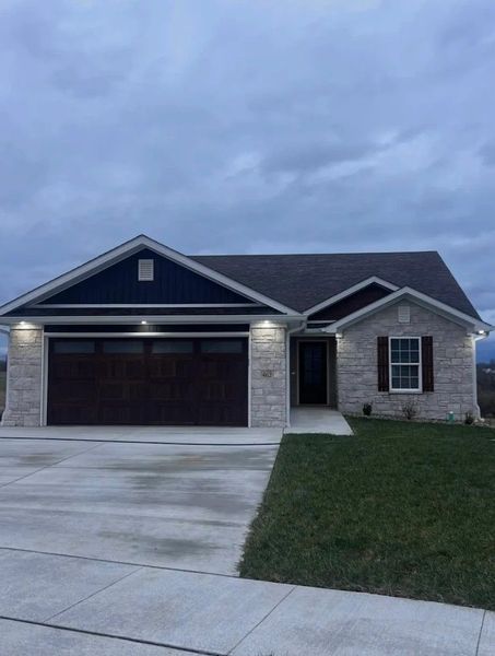 A single-story house with stone veneer, dark siding, a two-car garage, and a concrete driveway under a cloudy sky.