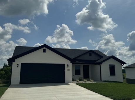 A single-story, white modern house with a dark roof and a two-car garage under a blue sky with fluffy clouds.