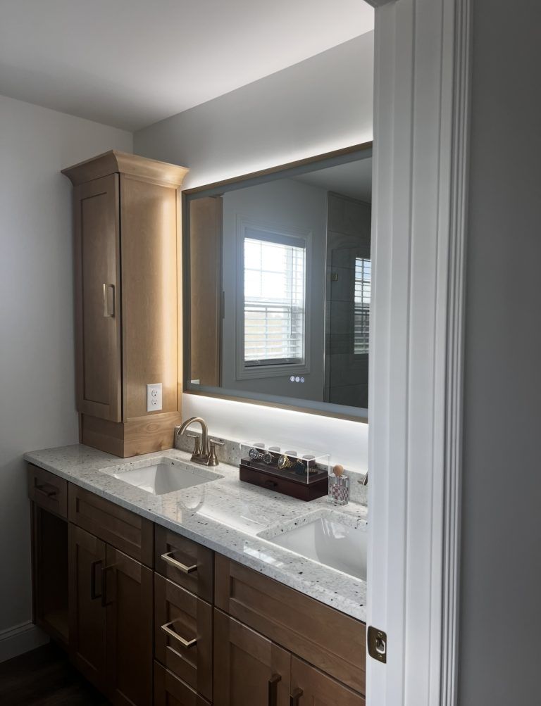 A modern double-sink bathroom vanity with light wood cabinets, speckled white countertops, and an illuminated mirror.