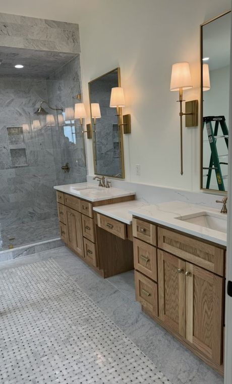 A bright, modern bathroom featuring double wood vanities, marble floors, a tiled walk-in shower, and brass wall sconces.
